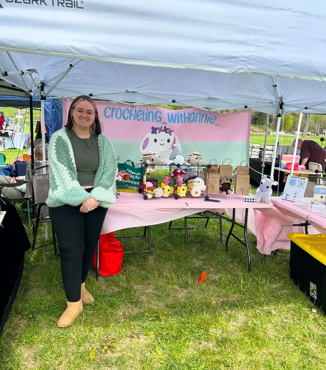 Annie smiling at her crochet market stall, surrounded by handmade plushies on a pink tablecloth